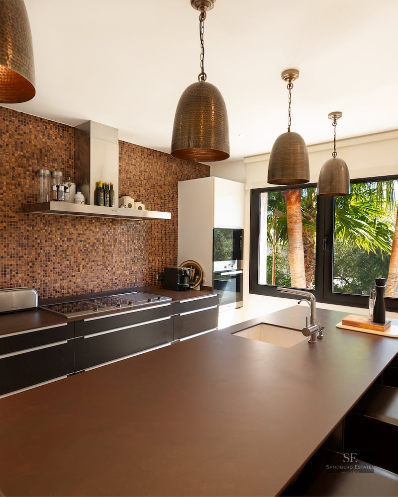 Modern kitchen featuring a large brown island, copper mosaic backsplash, and four bronze hammered pendant lamps.