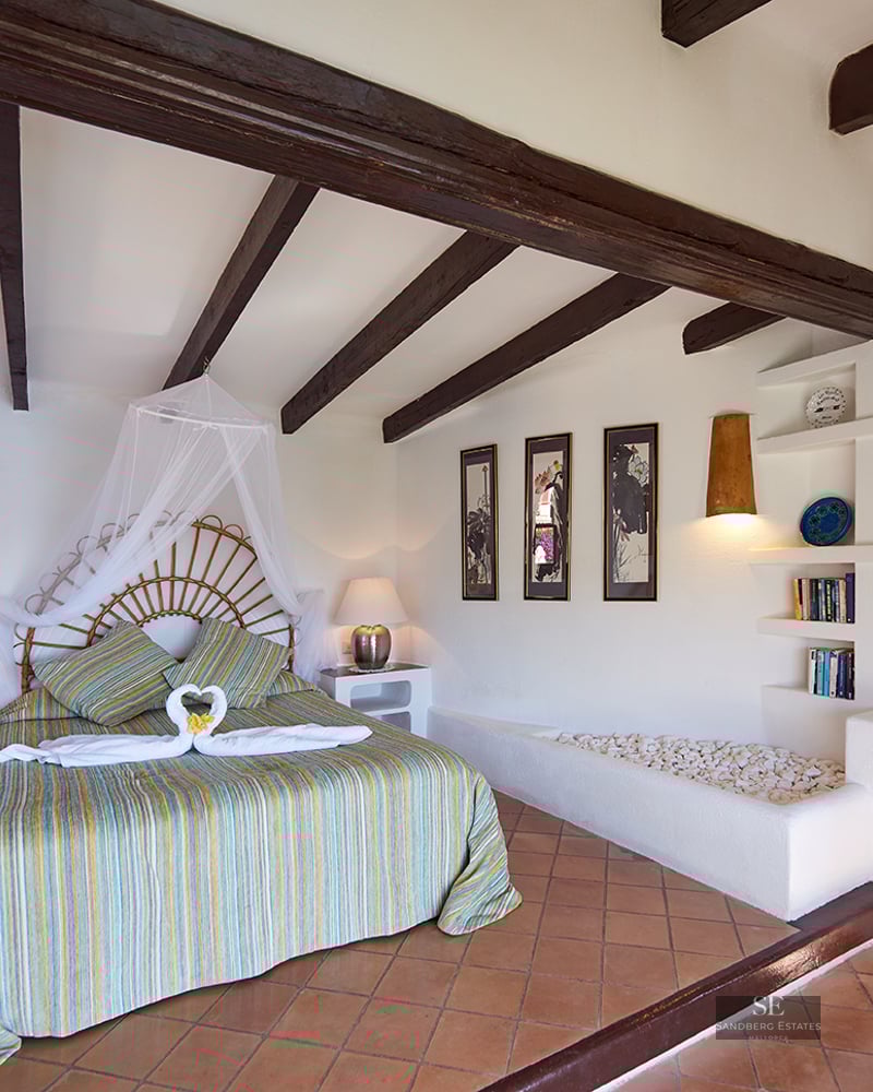 Bedroom with wicker headboard bed, dark wooden beams, terracotta floor, and white walls with built-in shelving.