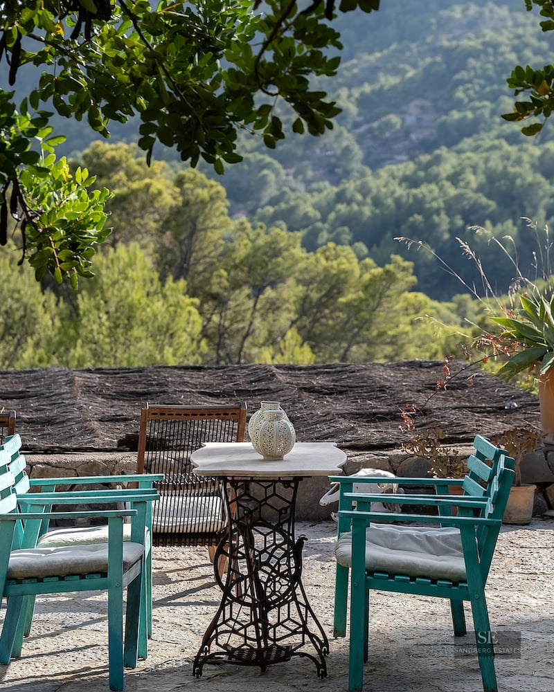Stone terrace with teal wooden chairs and a vintage table overlooking a lush green forest and mountain range.
