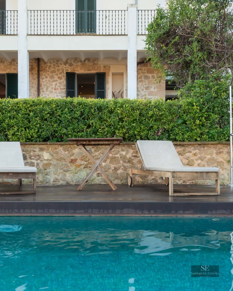 Turquoise swimming pool with wooden loungers in front of a stone Mediterranean house with green shutters.
