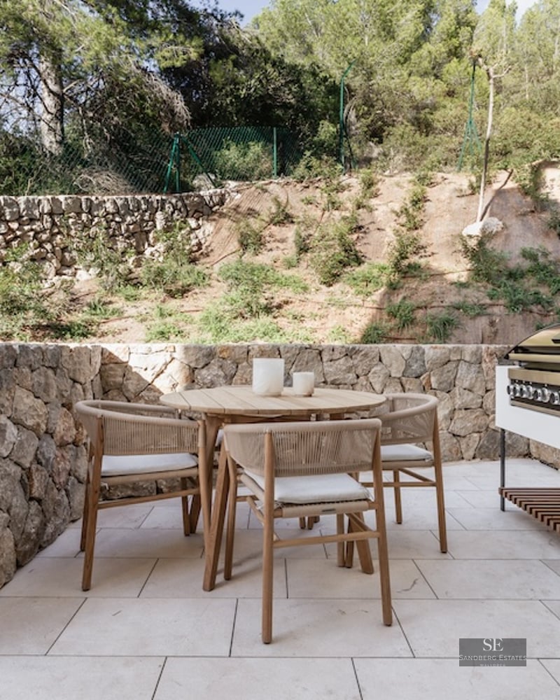 Outdoor terrace featuring a wooden dining set, stone walls, and a modern built-in grill area against a backdrop of trees.
