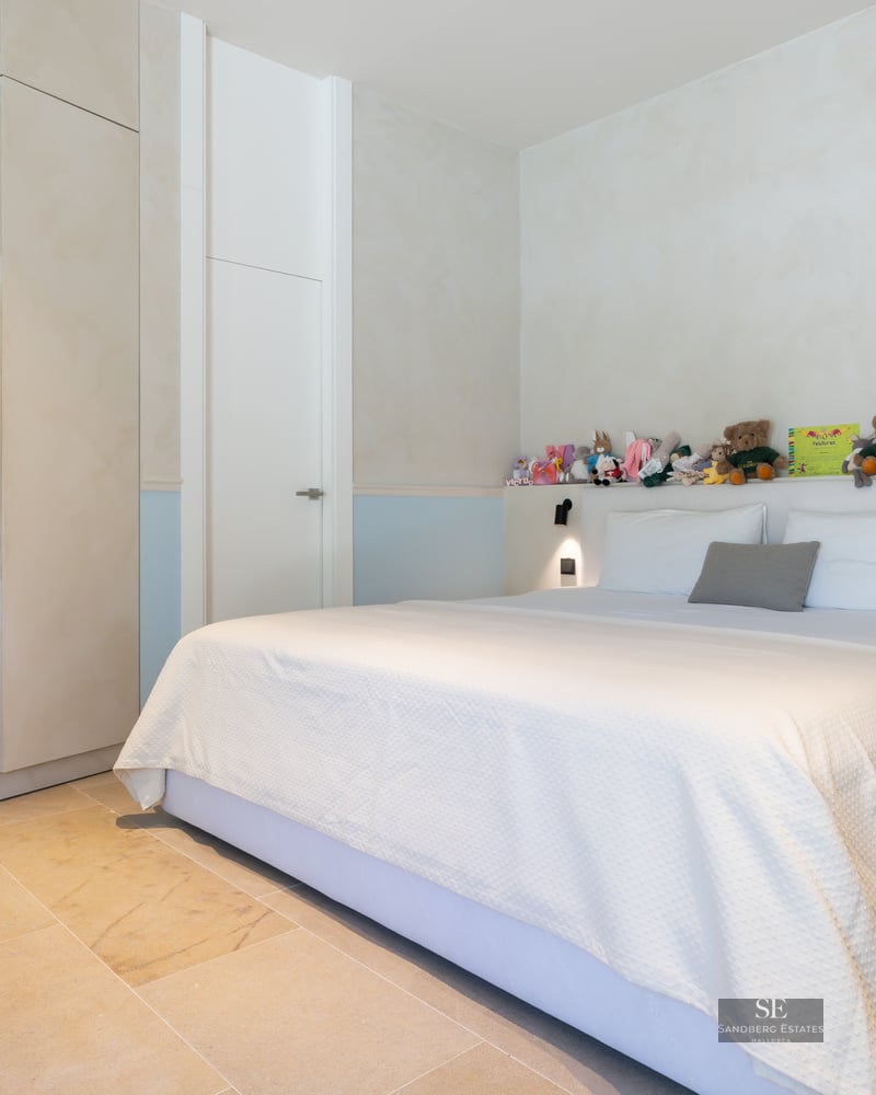 Bright bedroom featuring a large bed, stone floors, built-in wardrobes, and a row of stuffed animals on the headboard shelf.
