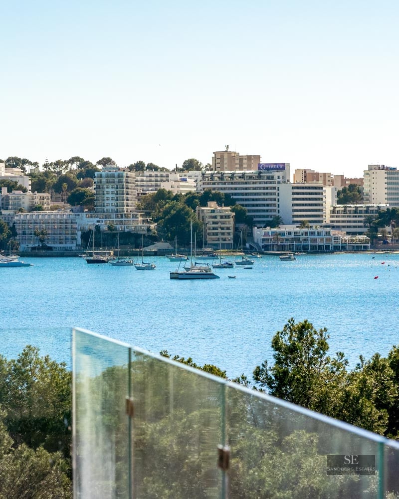 Glass balcony railing overlooking a bright blue bay filled with sailboats and a coastal city skyline.