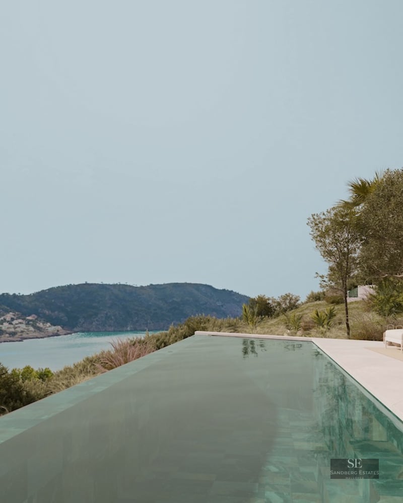 Infinity swimming pool on a stone terrace overlooking a turquoise bay and green hills under a clear sky.