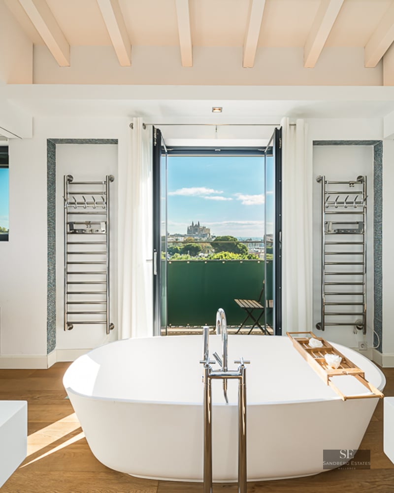 Freestanding white bathtub facing a balcony with city and cathedral views, featuring chrome towel rails.