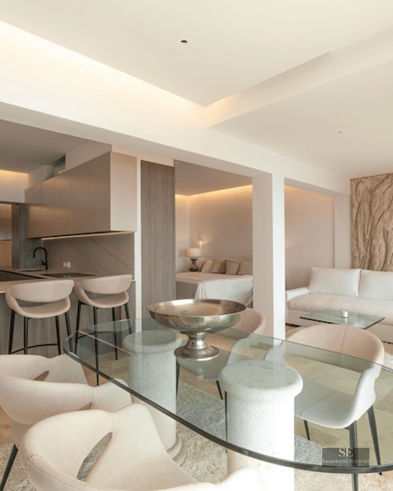 Modern open-plan living room featuring a glass dining table, cream sofa, and integrated kitchen in neutral stone tones.