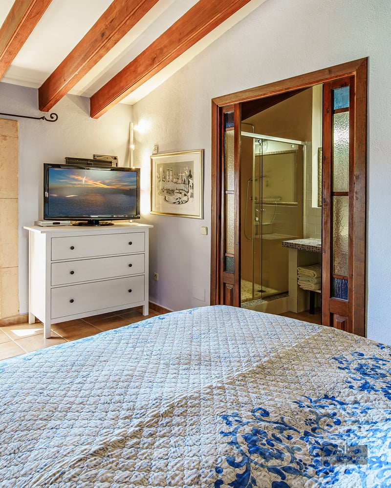 Bedroom with exposed wooden beams, a blue patterned bed, visible ensuite bathroom, and a stone-framed window.