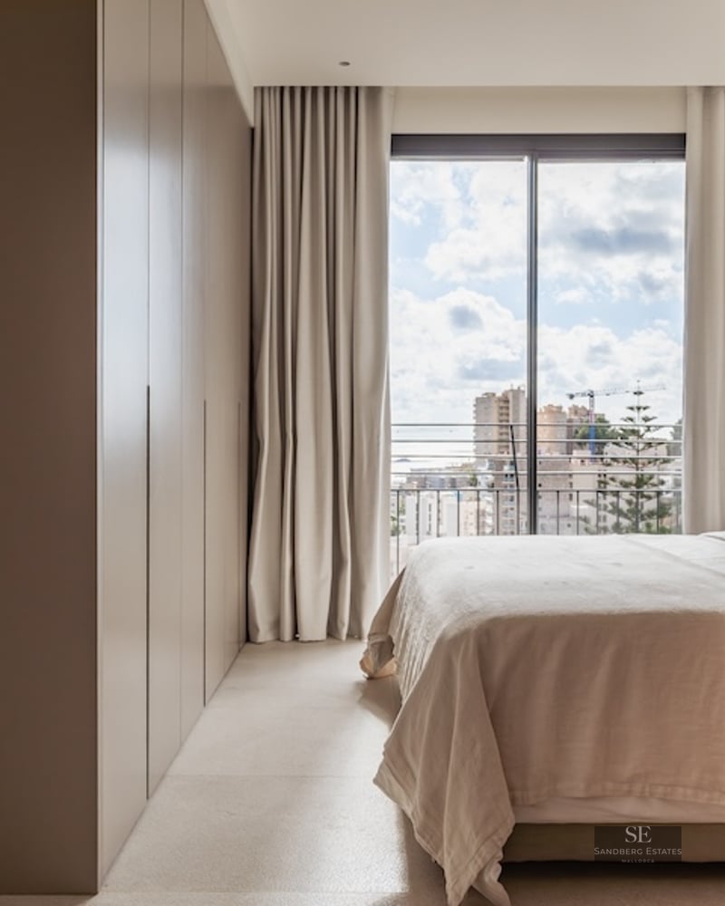 Bright bedroom with neutral tones, a large built-in wardrobe, and a floor-to-ceiling window overlooking a city.