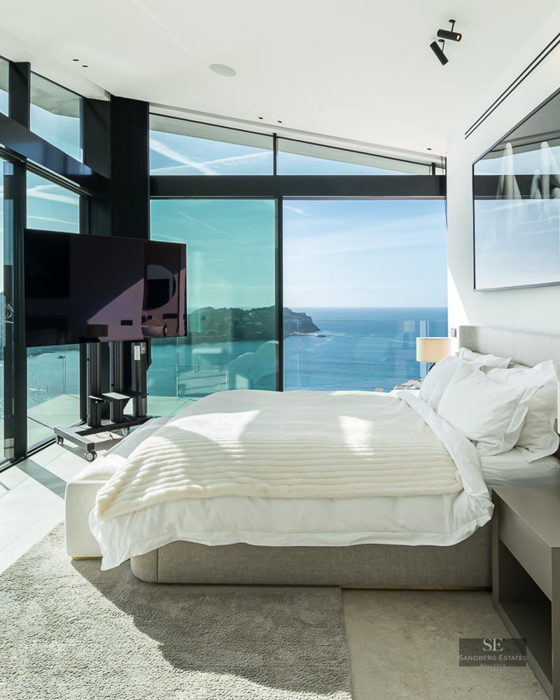 Bright master bedroom with floor-to-ceiling glass windows looking out over the ocean and a coastal town.