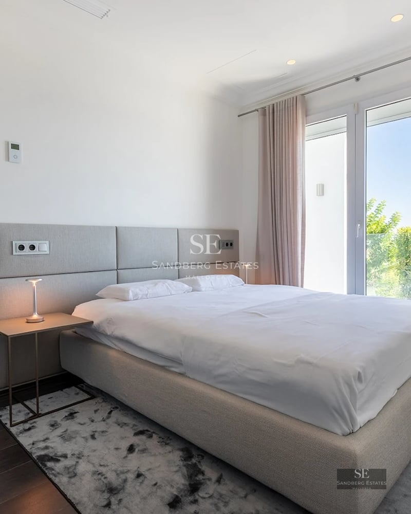 Minimalist bedroom with a large bed, ensuite bathroom view, and bright floor-to-ceiling windows with soft curtains.