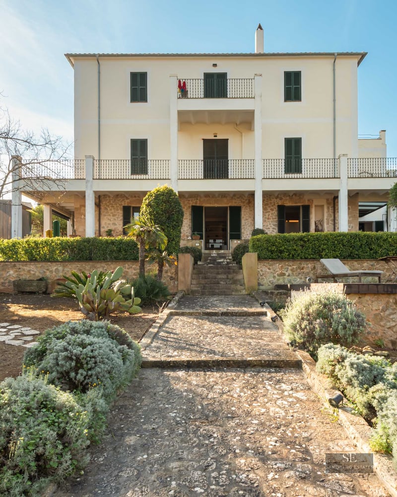 Rear view of a cream three-story villa with stone walls, green shutters, and a stone path through a garden.