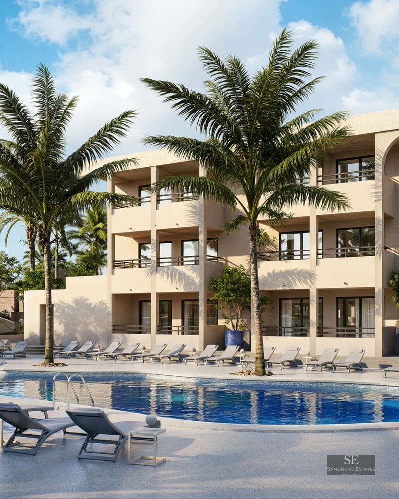 Grande piscine bleue avec chaises longues, palmiers et bâtiment méditerranéen moderne de trois étages sous un ciel clair.