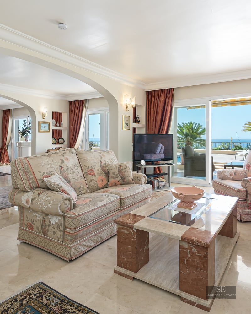 Bright living room with floral sofas, marble coffee table, and large glass doors opening to a terrace with sea view.