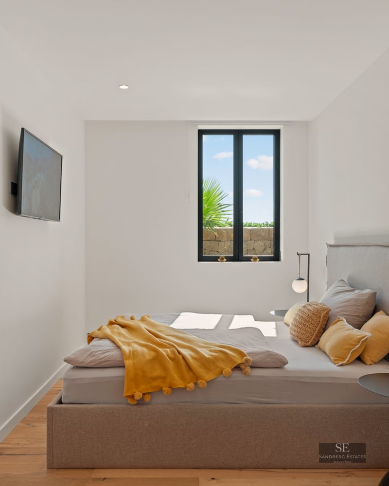 Bright bedroom featuring a taupe upholstered bed, yellow pom-pom blanket, minimalist lamps, and a scenic window view.