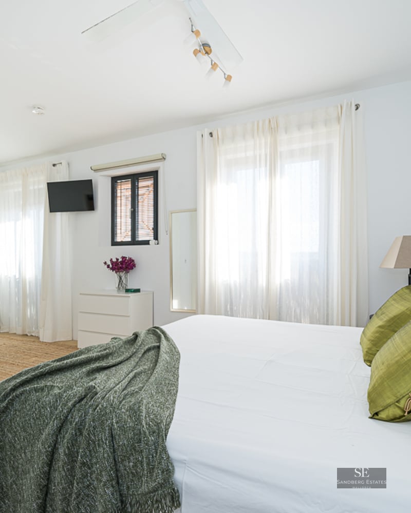 Bright master bedroom featuring a large bed with green pillows, a lounge area with a white sofa, and jute rugs.