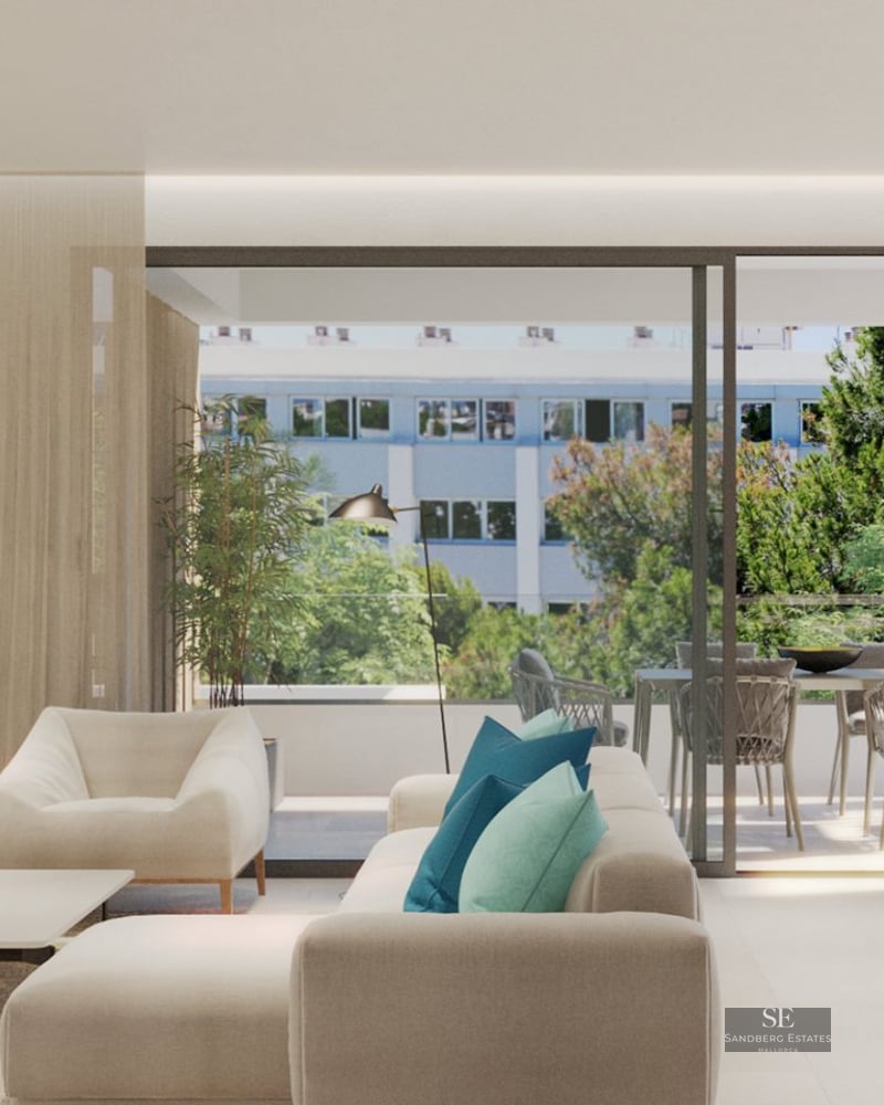 Bright living room with cream sofa and teal pillows opening to a sunlit balcony with trees in the background.