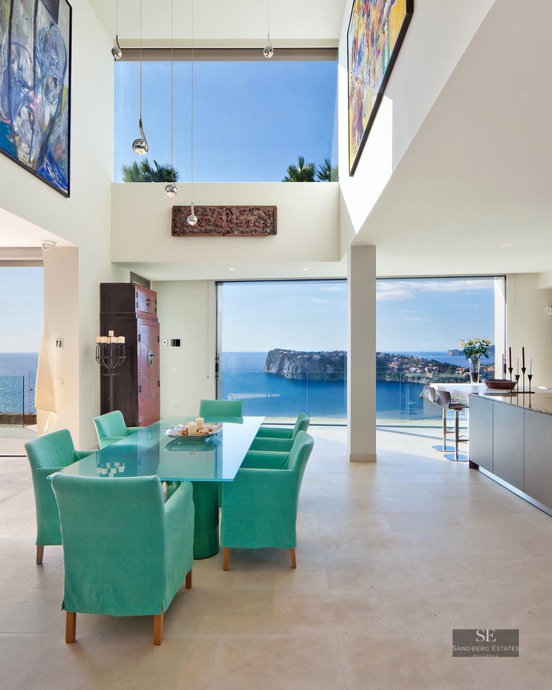 Modern open-plan dining and kitchen area with turquoise chairs and floor-to-ceiling windows overlooking the sea.