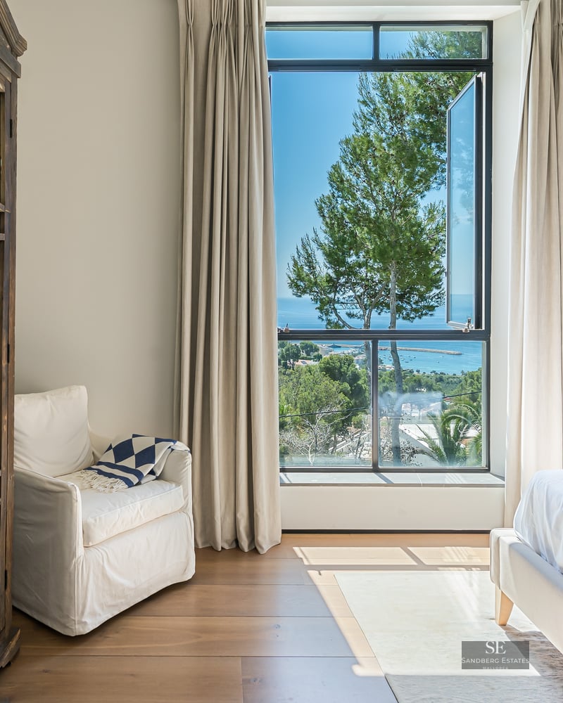 Bright master bedroom with large windows overlooking the sea, featuring a white armchair and ornate wooden wardrobe.