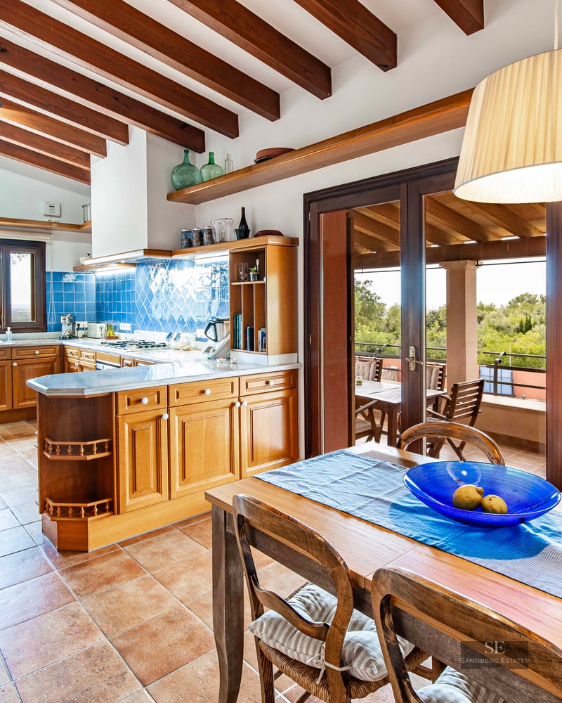 Kitchen with wood cabinets, blue tile backsplash, terracotta floor, and exposed wood beams with a dining table.