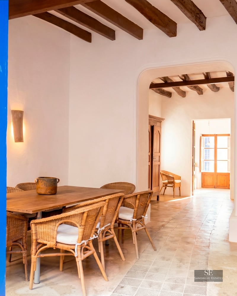 Dining area featuring a wooden table, wicker chairs, exposed ceiling beams, and a striking blue door in the foreground.