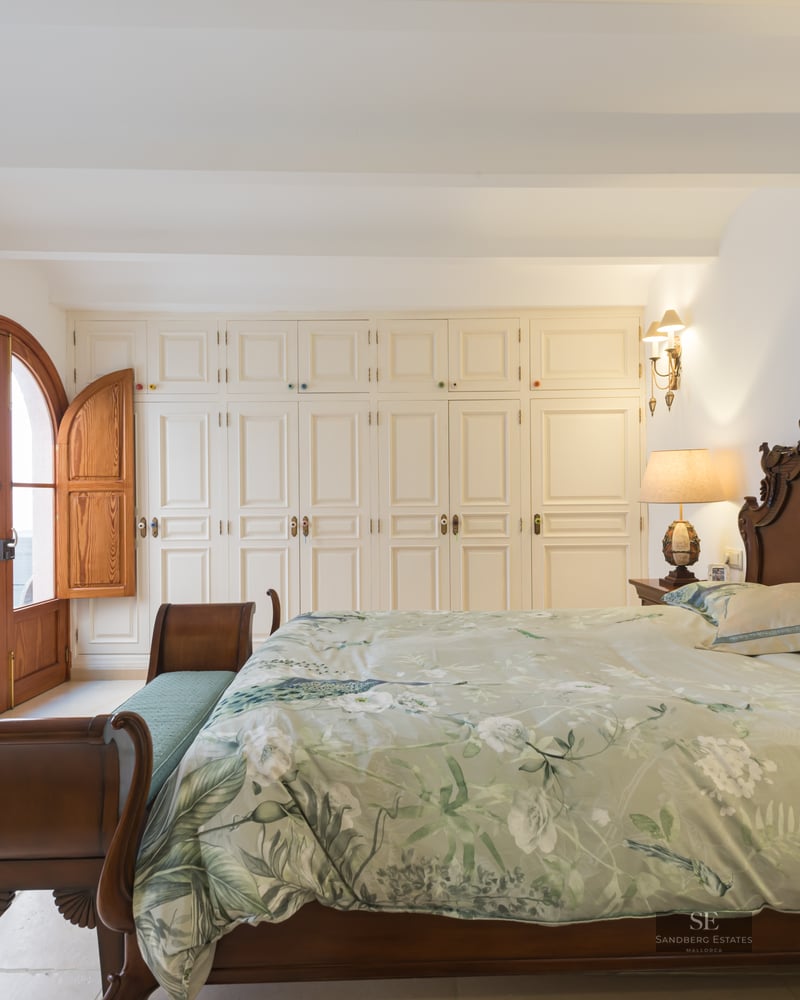 Spacious bedroom with a dark wood carved bed, floral bedding, white wardrobes, and arched wooden doors with shutters.