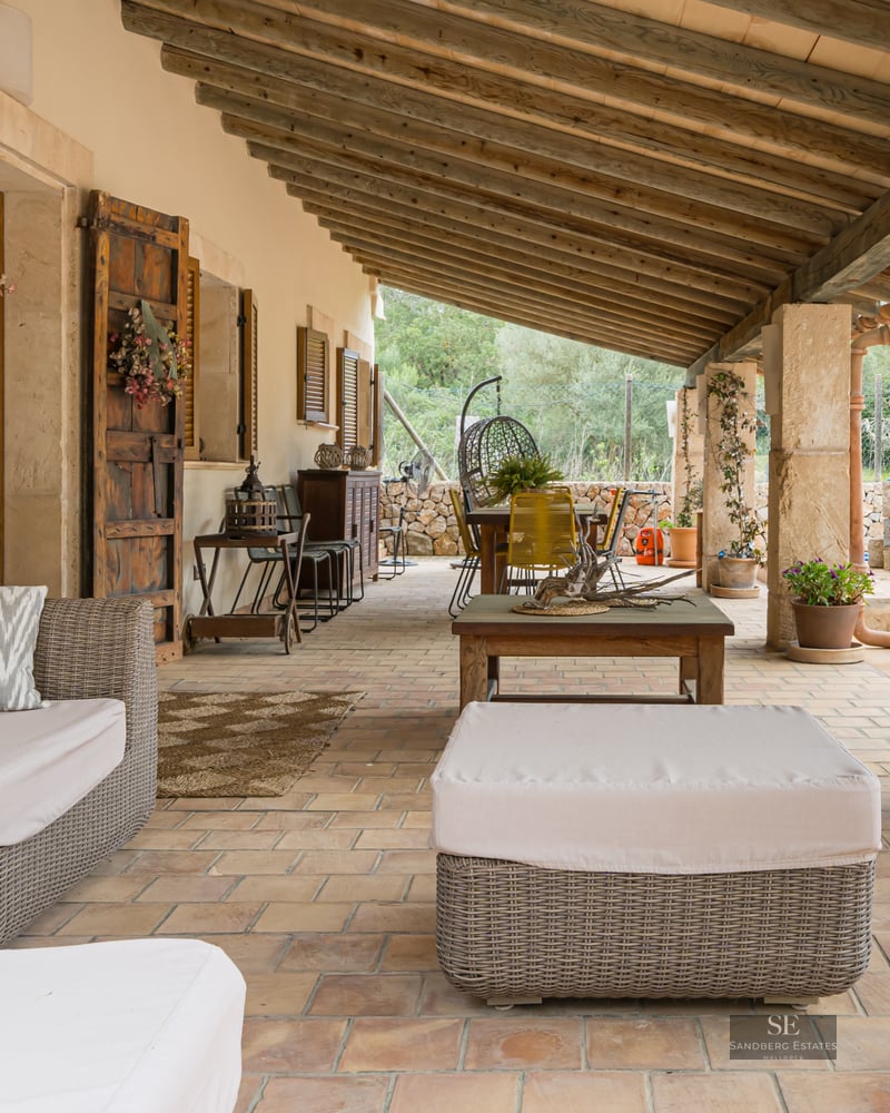 Shaded terrace featuring wicker sofas, exposed wooden beams, stone pillars, and terracotta tile flooring.