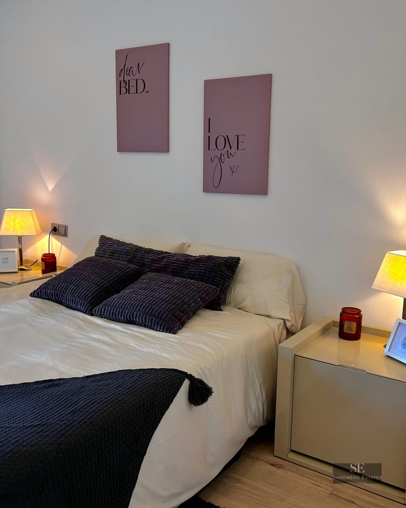 A cozy bedroom featuring a white bed with dark pillows, two bedside lamps glowing warmly, and pink wall art.