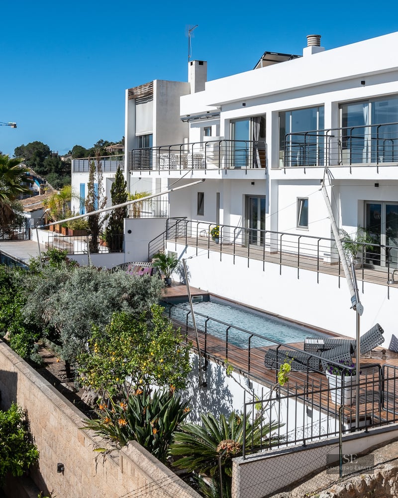 Modern white multi-level villa with a rectangular swimming pool, wooden decking, and lush Mediterranean landscaping.