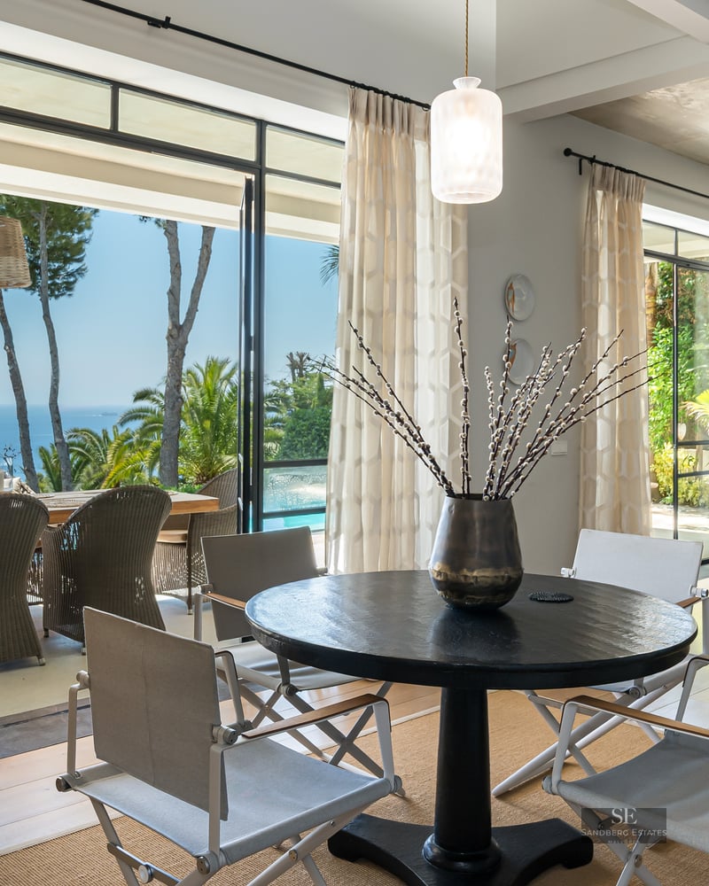 Modern dining room with a black round table and white chairs facing large glass doors with a view of the sea and terrace.
