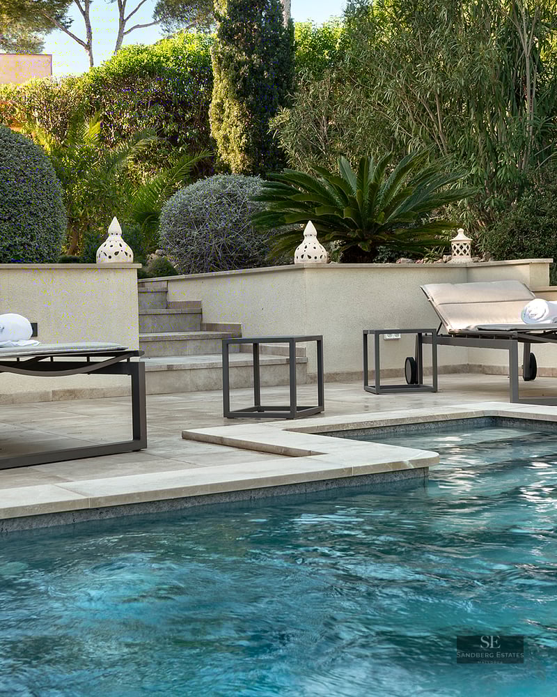 Blue swimming pool with modern sun loungers on a stone terrace surrounded by lush Mediterranean greenery.