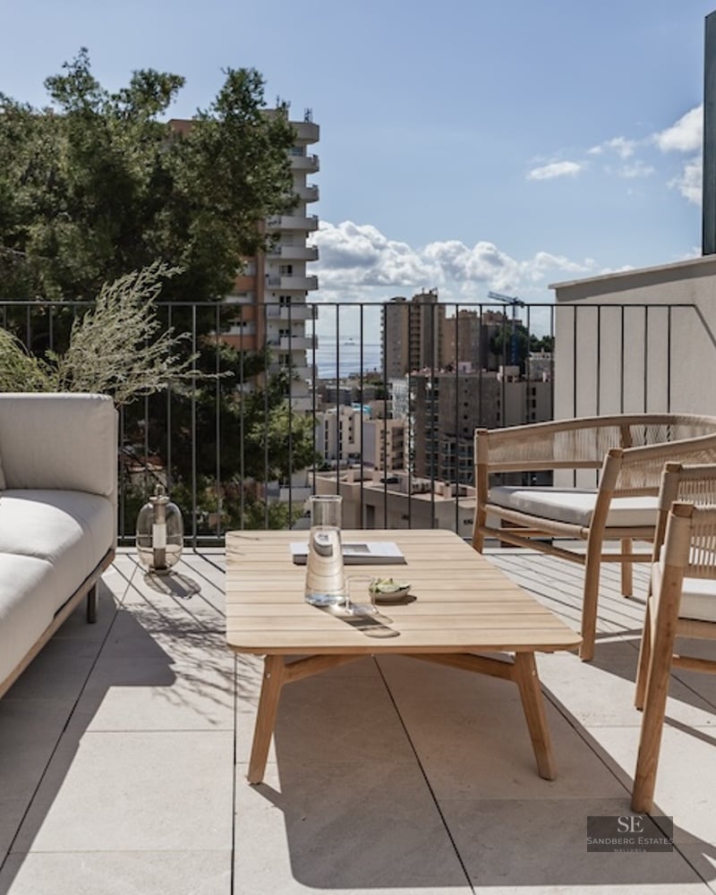 Luxury rooftop terrace with cream sofa, wooden armchairs, and a view of the city and sea under a bright sky.