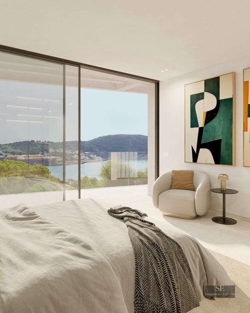 Modern master bedroom with large glass doors overlooking a coastal landscape, featuring abstract art and a lounge chair.