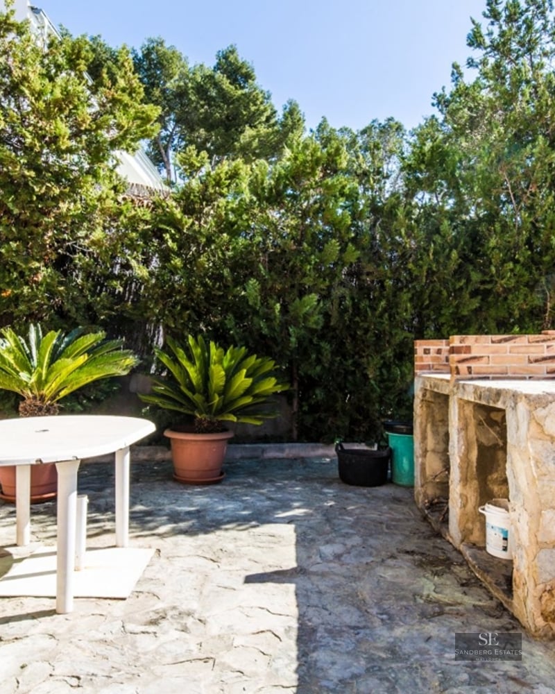 Paved patio with a stone barbecue, white outdoor table and bench, surrounded by lush green hedges and potted plants.