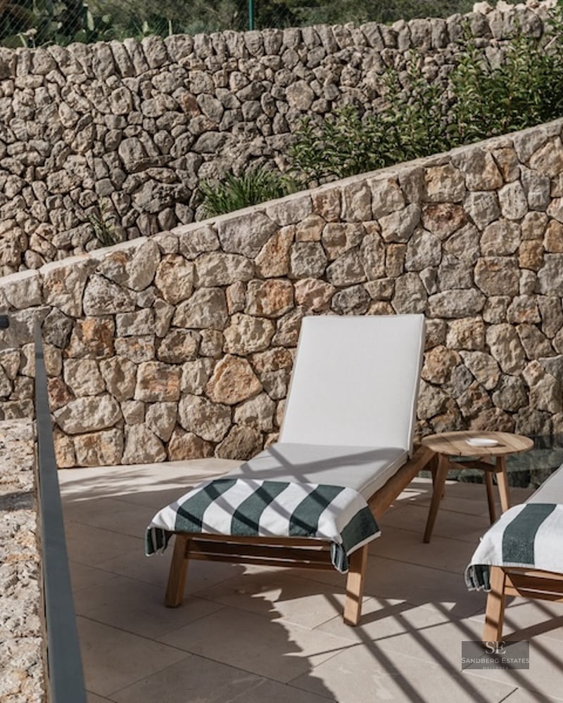 Two wooden sun loungers with white cushions in front of a rustic stone wall on a sunny terrace.