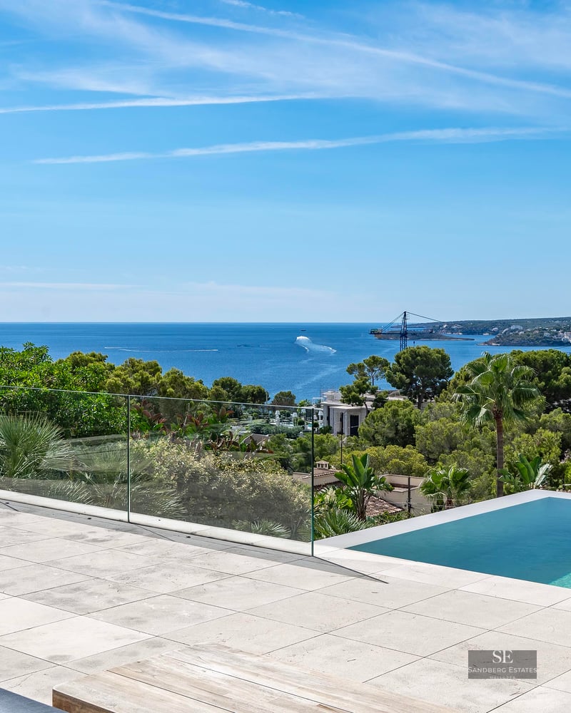 Modern stone terrace featuring an infinity pool with a glass railing overlooking the blue sea and lush greenery.