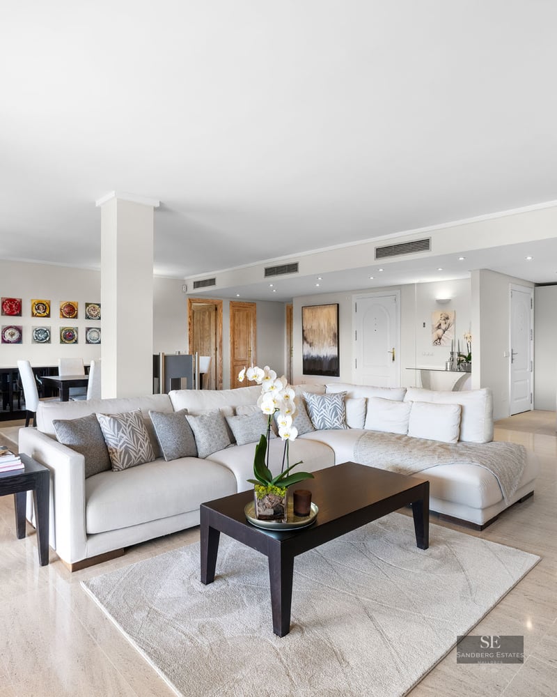 Spacieux salon avec canapé en L blanc, table basse en bois sombre, sol en marbre et cuisine intégrée.