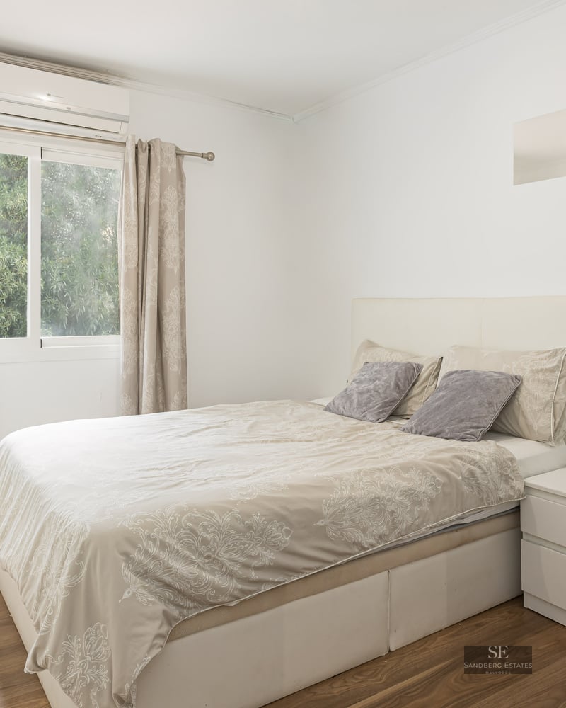 Chambre lumineuse avec lit double, meubles blancs, parquet et fenêtre donnant sur des arbres.