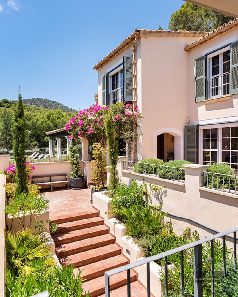 Sunny peach-colored villa with green shutters, terracotta stairs, and lush garden overlooking a forested valley.