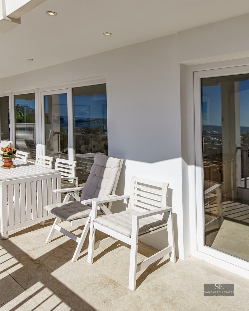 Sunlit terrace with white outdoor dining set and large glass sliding doors reflecting the coastline.