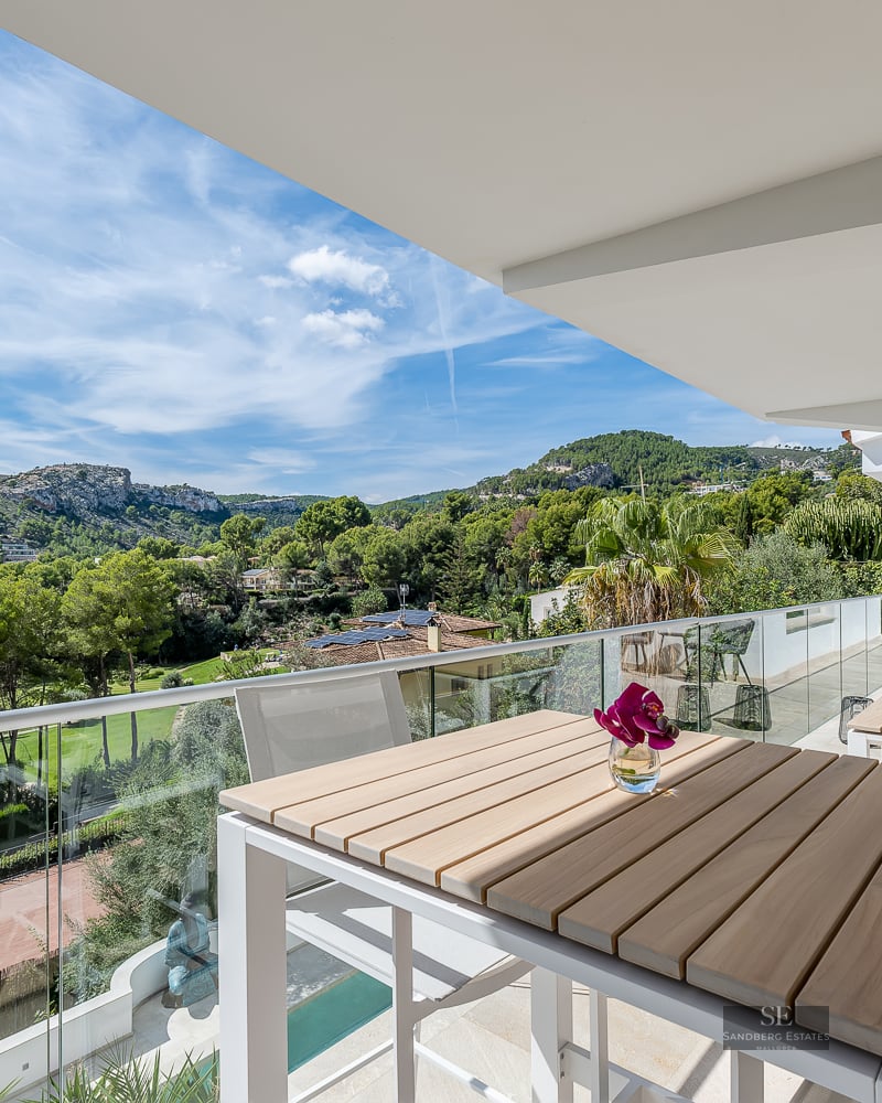 Modern balcony with wooden table and glass railing overlooking a lush green valley and mountains under a blue sky.