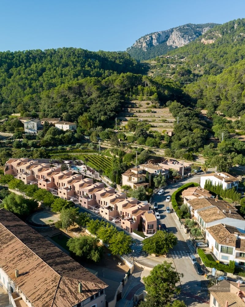 Aerial drone shot showing modern terracotta houses in a Mediterranean village surrounded by forested mountains.