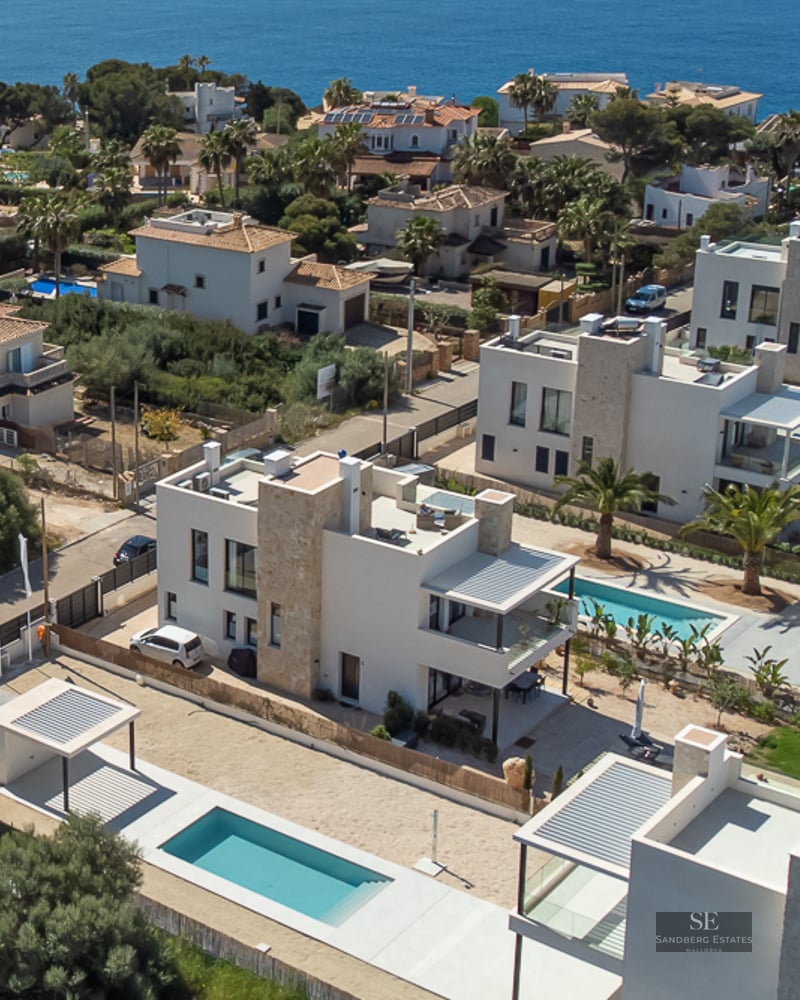 Drone view of modern white villas with private pools and stone accents in a coastal Mediterranean neighborhood.