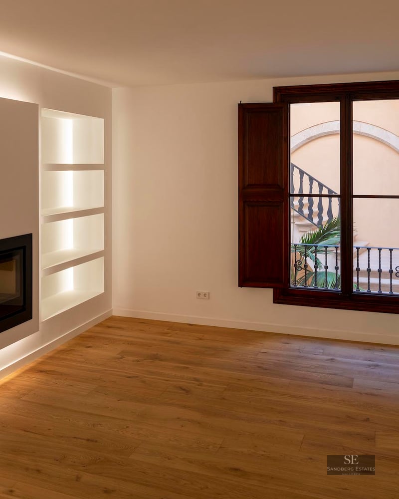 Modern empty room featuring a built-in fireplace, backlit shelving, oak floors, and dark wood shutters looking out to a courtyard.