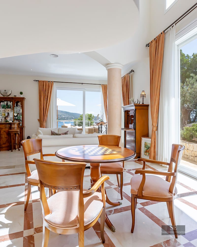 Spacious living area with marble floors, wooden table, pillars, and large windows overlooking a garden and sea.