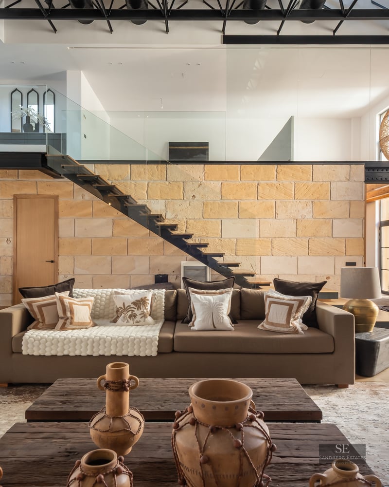 Modern living room featuring a stone wall, floating staircase, large wicker lamps, and a comfortable brown sofa.