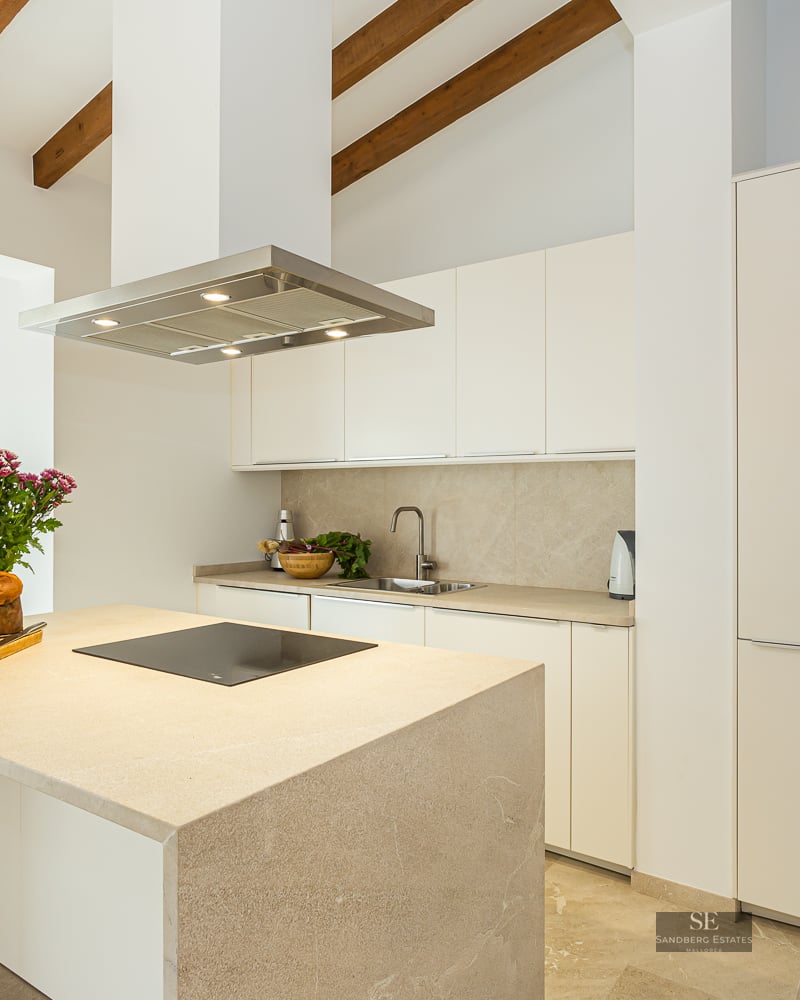 Bright kitchen featuring a stone island, exposed wooden ceiling beams, and minimalist white cabinetry.
