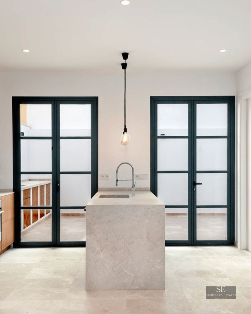 Minimalist kitchen featuring a central beige stone island, walnut wood cabinetry, and large black-framed glass doors.