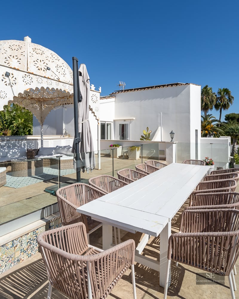 Spacious terrace with a white dining table, wicker chairs, BBQ station, and an ornate white gazebo under a clear blue sky.