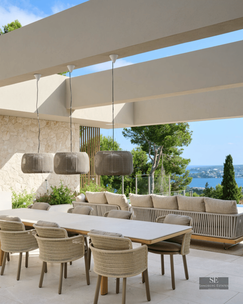 Modern terrace with a long dining table, outdoor kitchen, and sofas overlooking the Mediterranean sea and coastline.