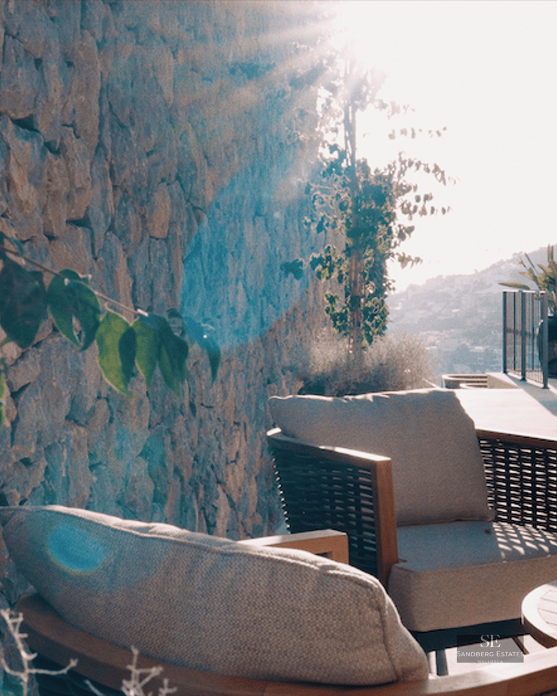 Sun-drenched terrace featuring elegant grey armchairs, a wooden table, a rustic stone wall, and white arched pillars.